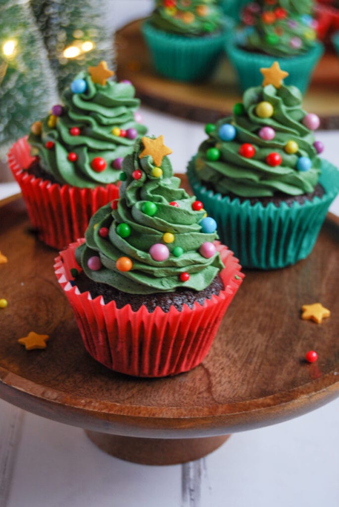 chocolate cupcakes in red and green wrappers decorated with green buttercream frosting, coloured sprinkles and a gold star to resemble Christmas trees on a wooden cake stand.