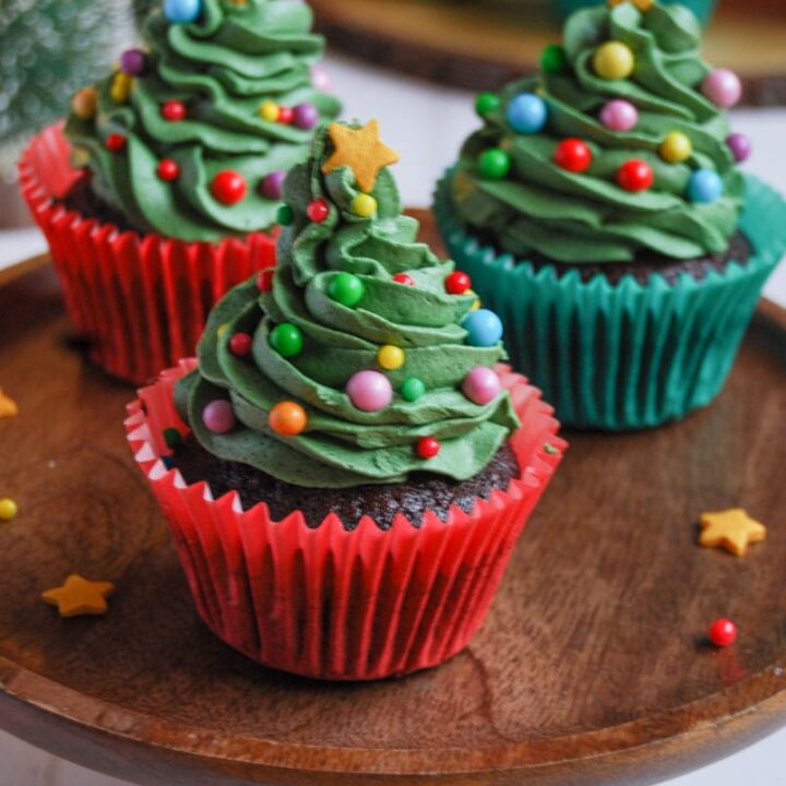 chocolate cupcakes in red and green wrappers decorated with green buttercream frosting, coloured sprinkles and a gold star to resemble Christmas trees on a wooden cake stand.