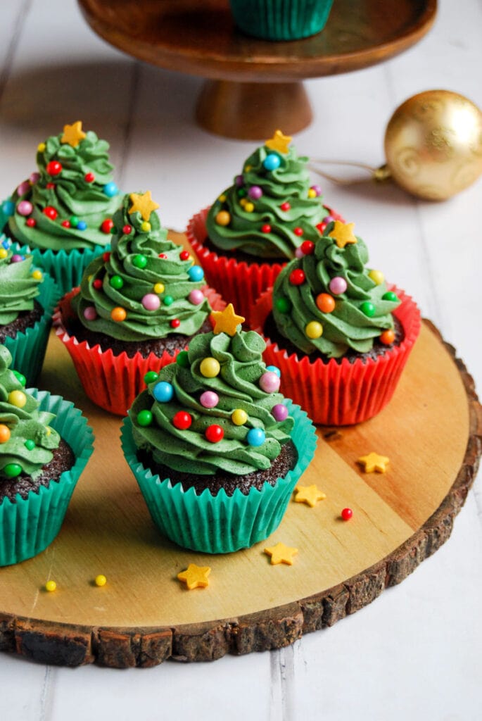 Chocolate cupcakes decorated with green buttercream icing, coloured round sprinkles and a gold star to resemble a Christmas tree on a wooden serving board.