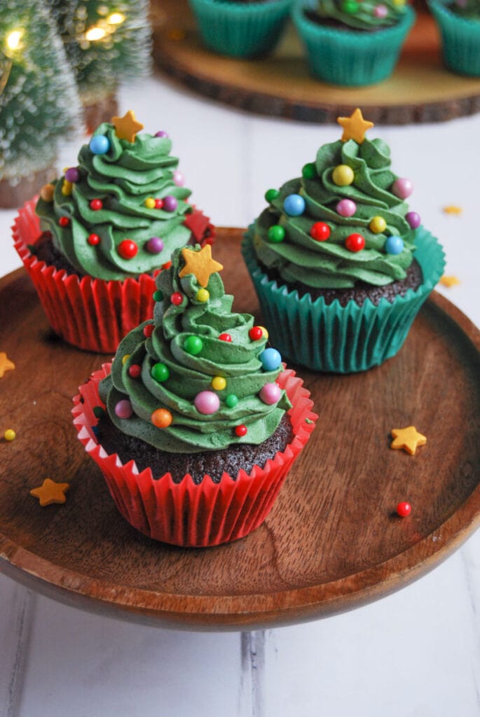 Chocolate cupcakes decorated with green buttercream frosting, multi coloured sprinkles and a gold star to resemble Christmas trees on a wooden cake stand.