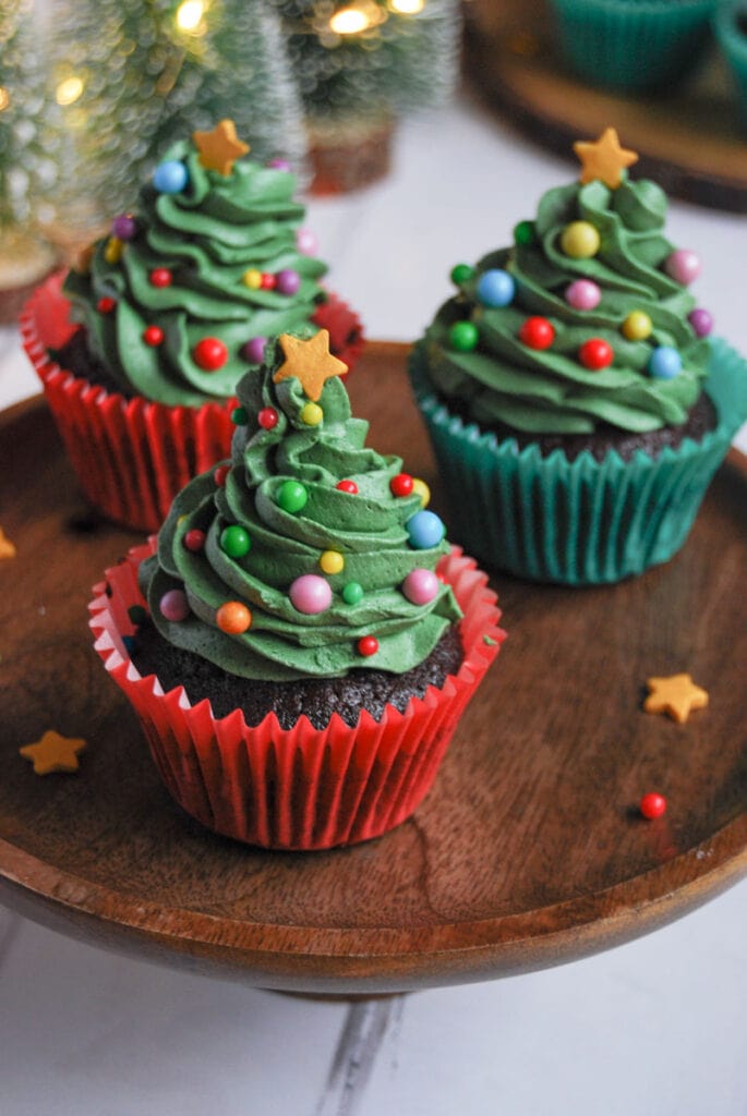 three Christmas cupcakes decorated with green buttercream frosting, coloured sprinkles and a gold star on a wooden cake stand.