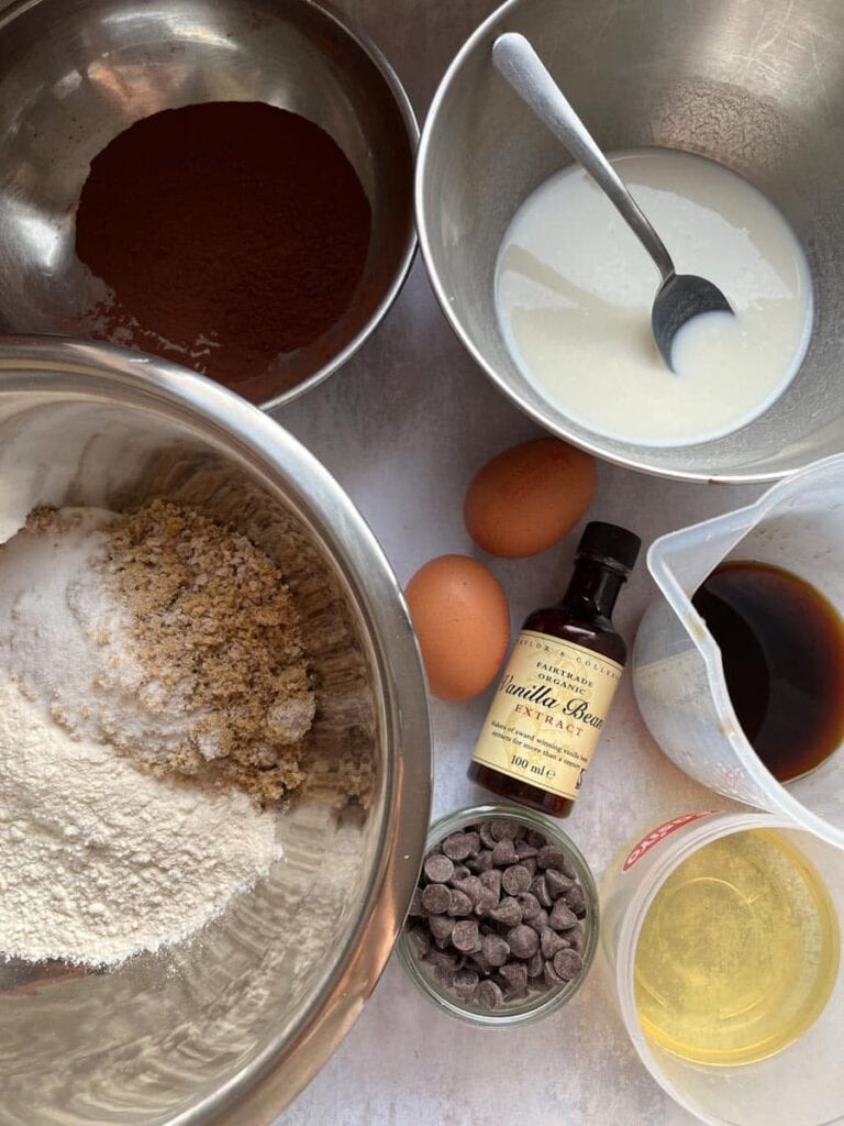 a large silver mixing bowl of flour and brown sugar, a jar of chocolate chips, two eggs, a bottle of vanilla extract, bowls of cocoa powder and buttermilk and a jug of cold coffee.