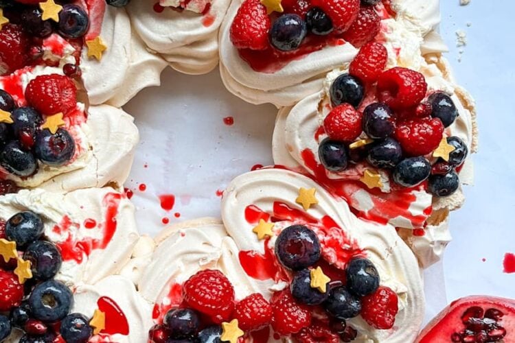 a pavlova in a wreath shape topped with whipped cream, raspberries, blueberries, gold stars and raspberry coulis and half a pomegranate on a white backdrop.