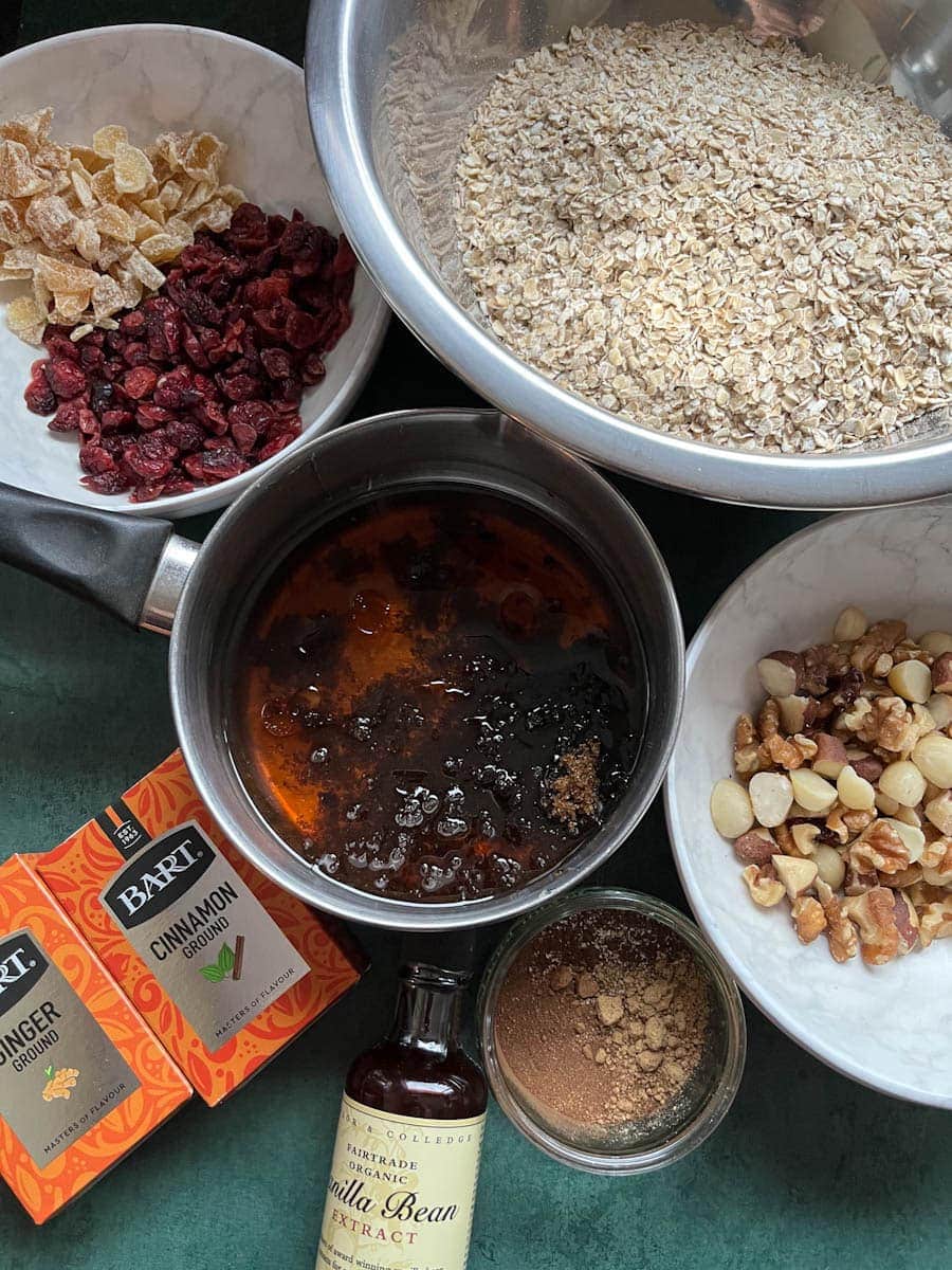 a large bowl of oats, a pan of honey and brown sugar, white bowls of mixed nuts, dried cranberries and candied ginger, a bottle of vanilla extract and packets of ground cinnamon and ginger.