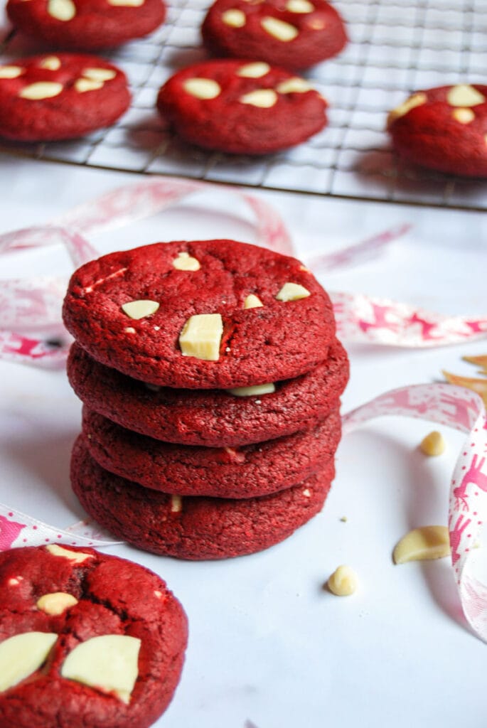 four red velvet cookies with white chocolate chunks stacked on top of each other with red Christmas ribbon. More cookies can be seen in the background on a wire rack.