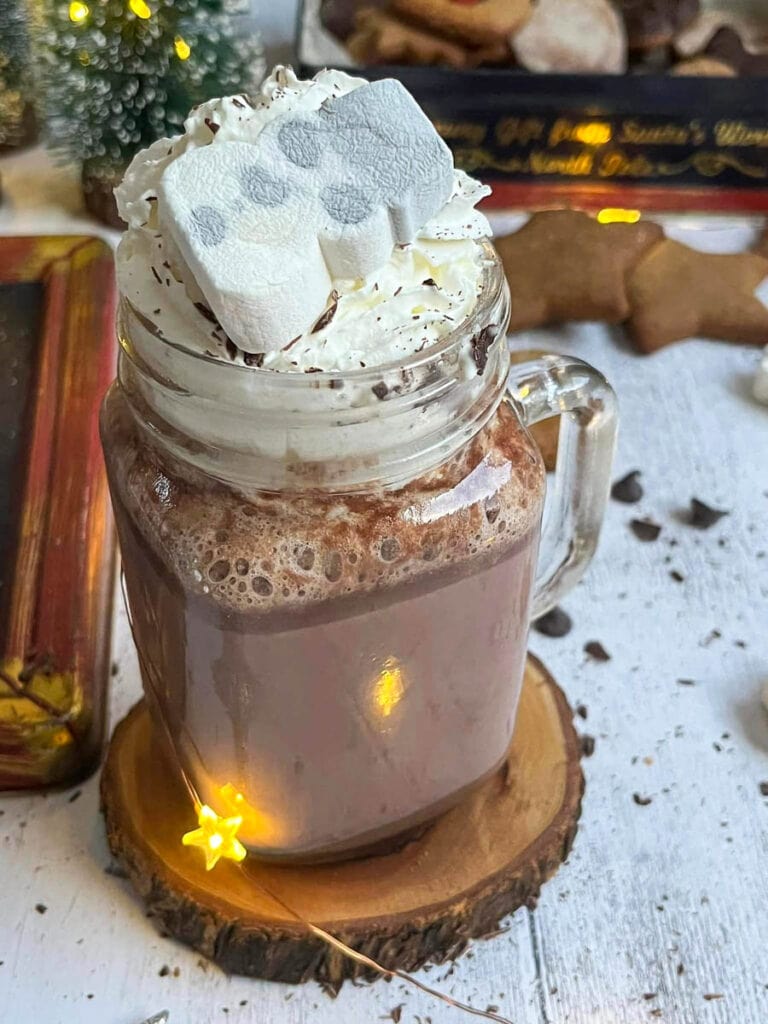 a glass mug of hot chocolate with whipped cream and a marshmallow snowman on a wooden coaster with star lights and gingerbread star cookies in the background.