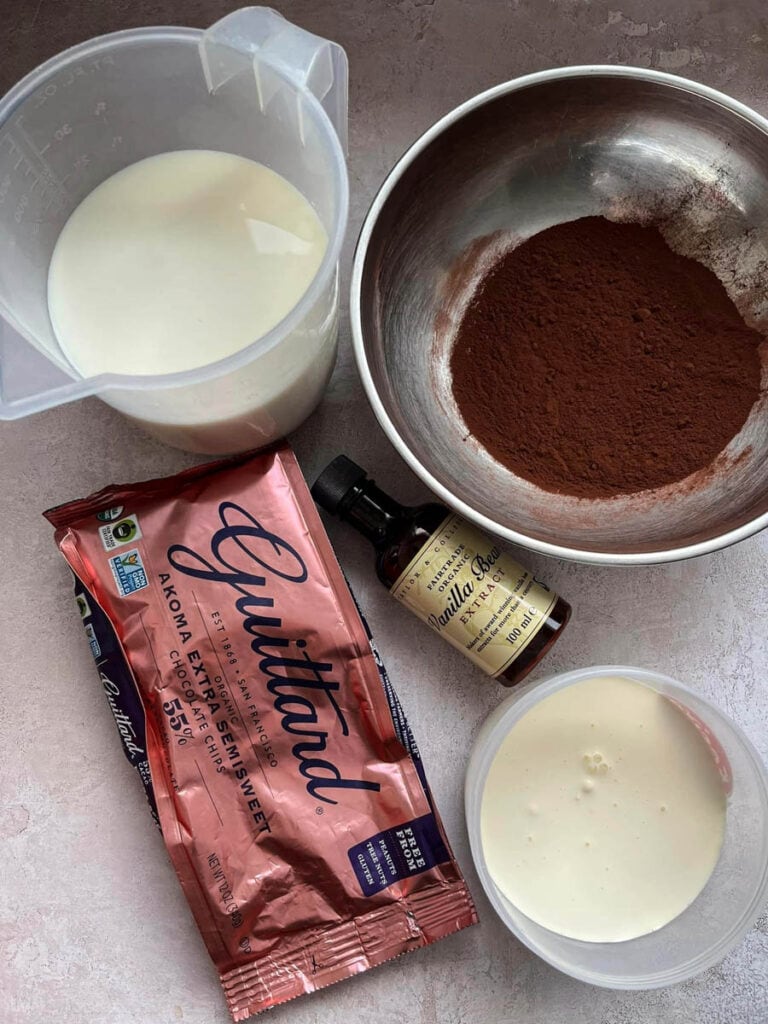 a packet of chocolate chips, a silver bowl of cocoa powder, a bottle of vanilla extract, a tub of double cream and a jug of milk.