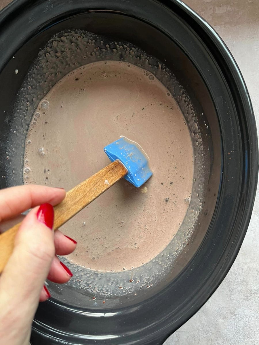 milk, cream and chocolate chips being stirred with a blue wooden handled spatula in a slow cooker bowl.