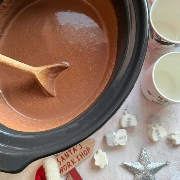 Hot chocolate in a slow cooker bowl with a wooden spoon, santa and snowflake marshmallows, Christmas mugs and a Santa's workshop sign.