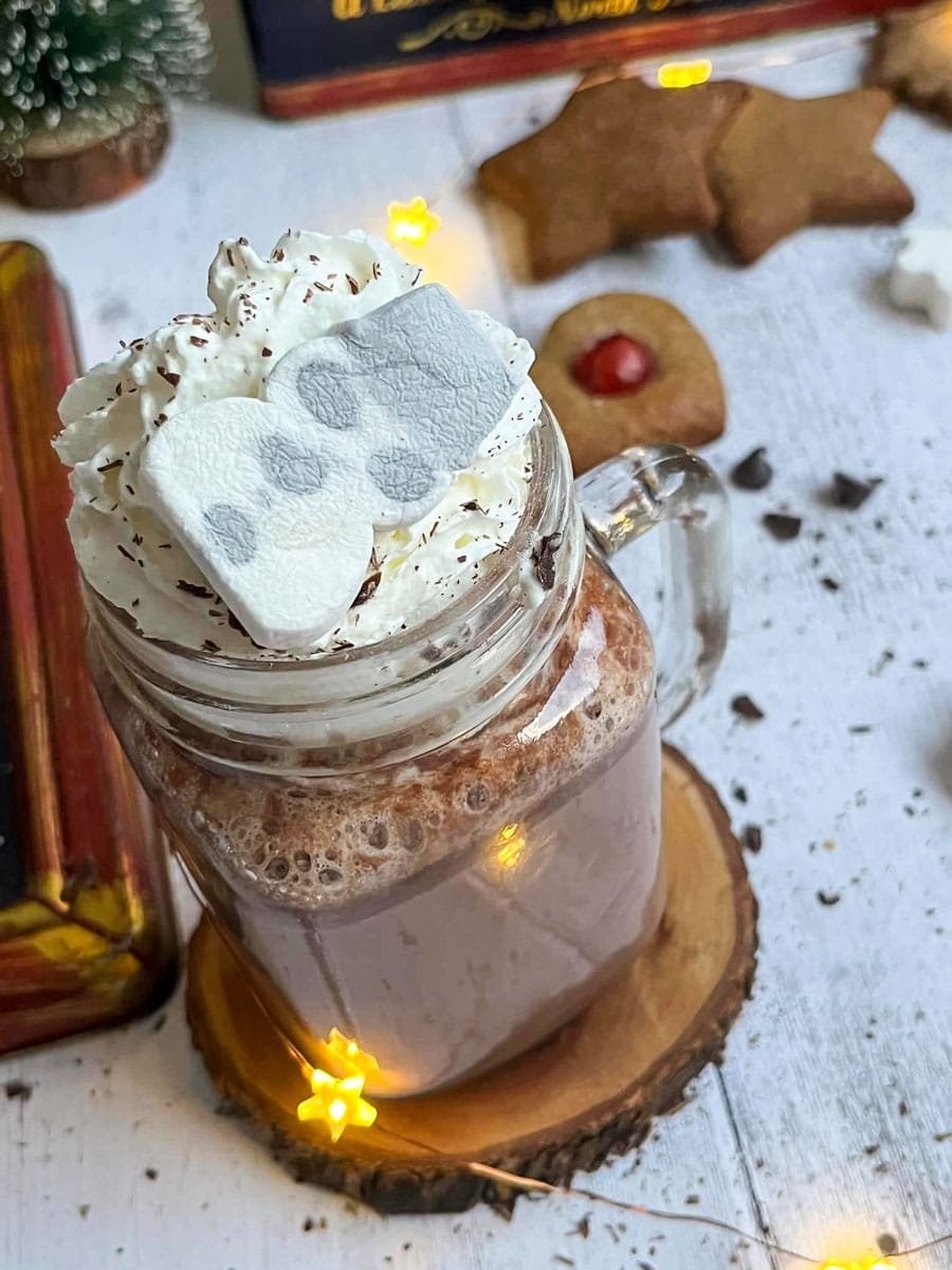 a glass mug of hot chocolate topped with whipped cream, grated chocolate and a snowman marshmallow.