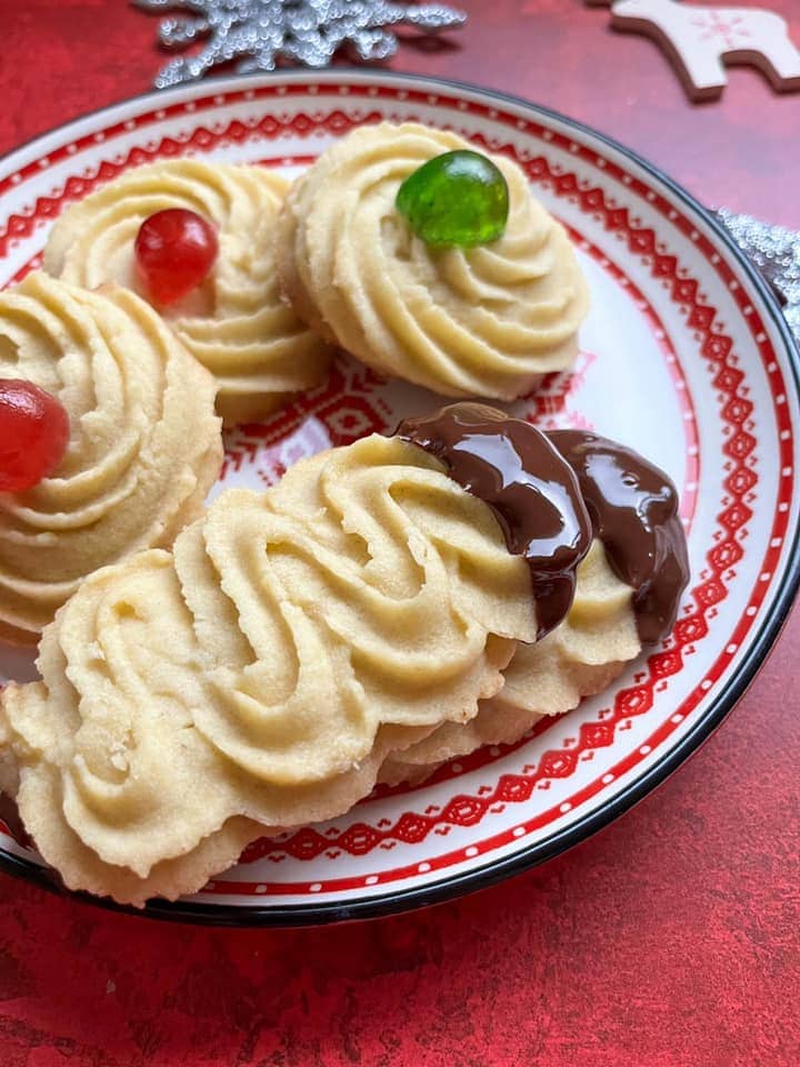 two chocolate dipped Viennese biscuits and three round biscuits topped with red and green cherries on a red and white plate.