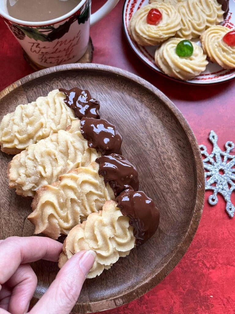 chocolate dipped piped long biscuits on a wooden cake stand, a mug of coffee and a plate of round piped biscuits tipped with red and green glace cherries.