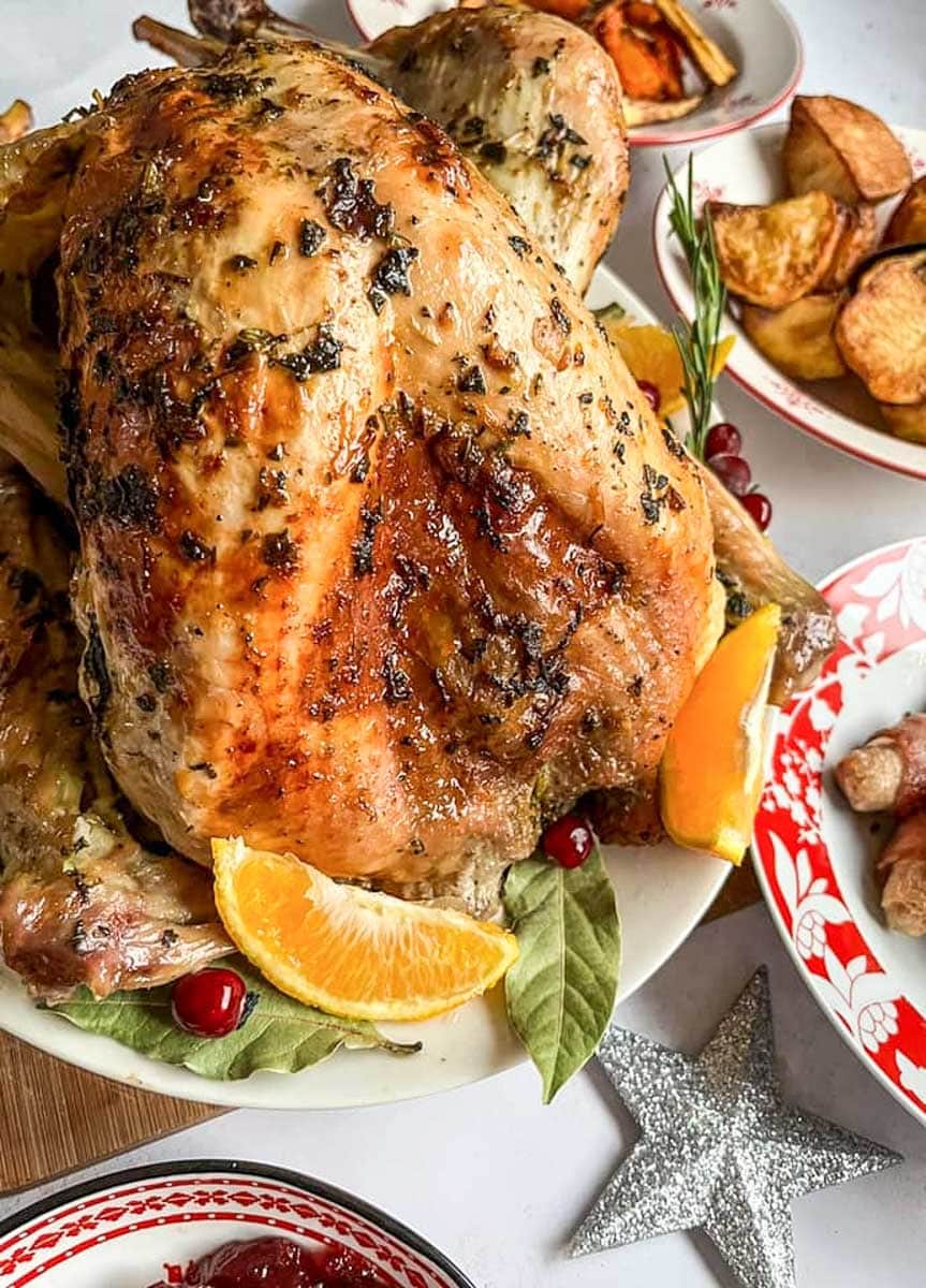 A roasted turkey on a white plate with orange slices, bay leaves, rosemary sprigs and cranberries and a bowl of roast potatoes.