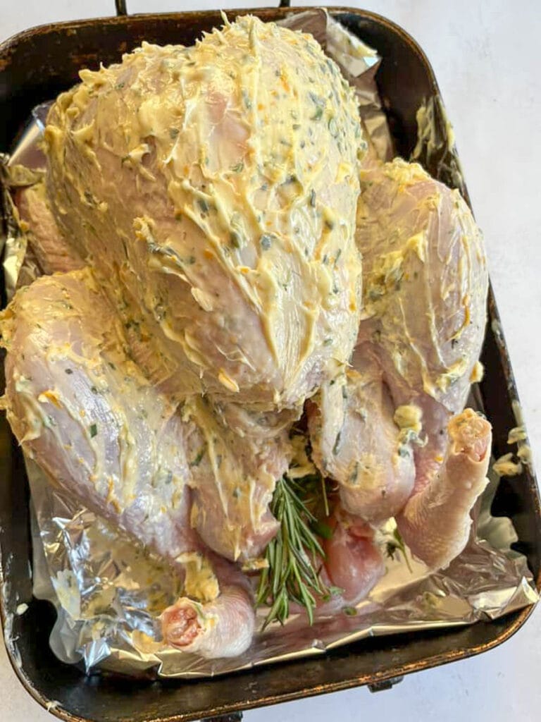 an uncooked large turkey covered in butter and stuffed with fresh rosemary and thyme in a foil lined roasting tin.