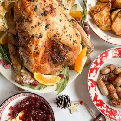 a roast turkey on a large serving pate with orange slices, cranberries, bay leaves and fresh rosemary, a dish of cranberry sauce and red and white dishes of roast potatoes and bacon wrapped sausages.