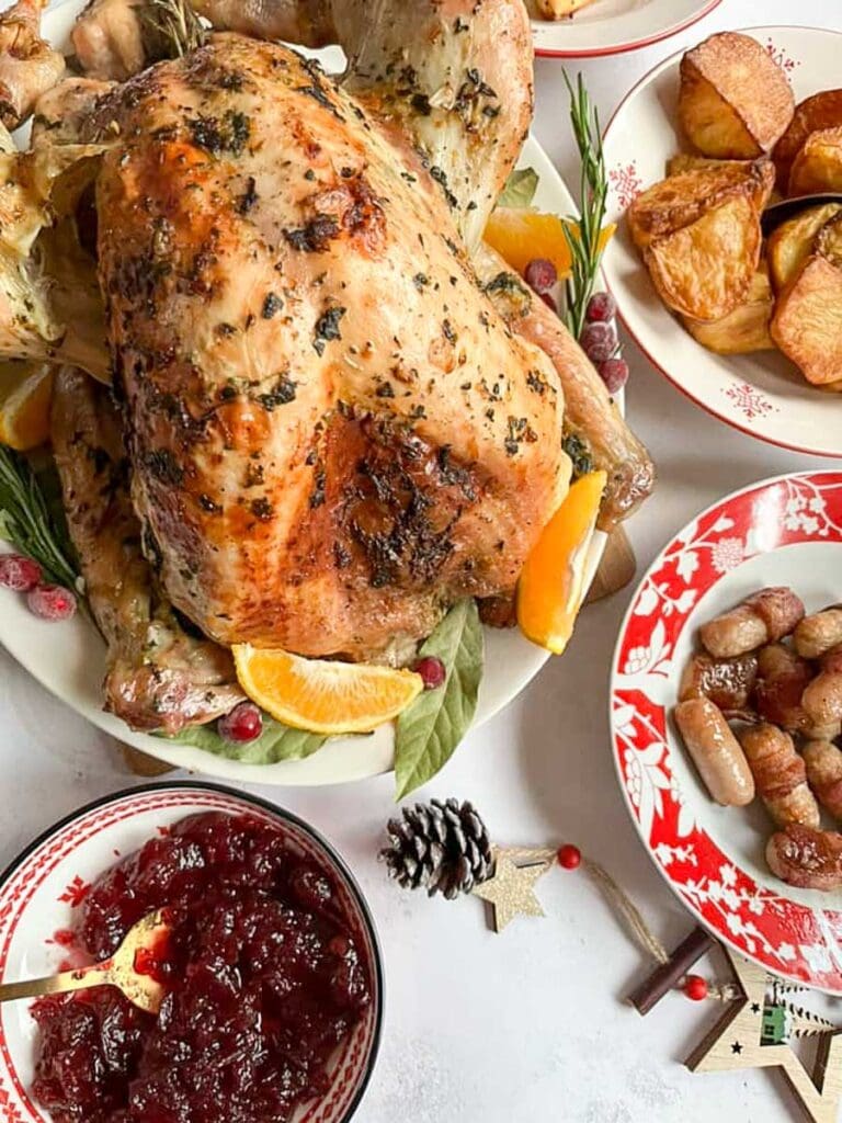 a roast turkey on a large serving pate with orange slices, cranberries, bay leaves and fresh rosemary, a dish of cranberry sauce and red and white dishes of roast potatoes and bacon wrapped sausages.