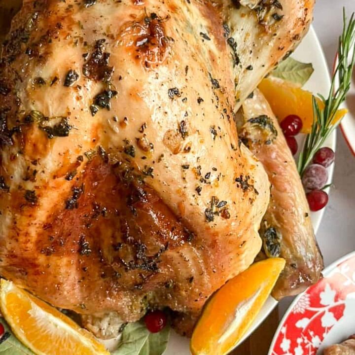 a roast turkey with herb butter on a white serving plate with orange slices, bay leaves, fresh rosemary and cranberries.