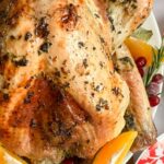 a roast turkey with herb butter on a white serving plate with orange slices, bay leaves, fresh rosemary and cranberries.