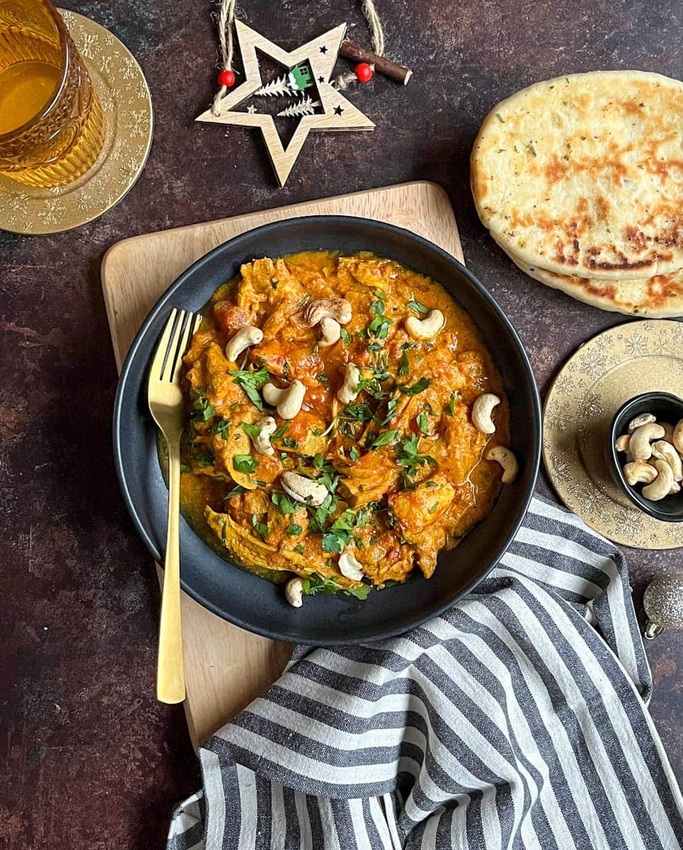 a black stoneware dish of turkey curry with freshly chopped coriander and cashew nuts with a gold fork. A grey and white striped tea towel, naan bread, a glass of water, a small black dish of cashew nuts on a gold coaster and a wooden Christmas decoration.