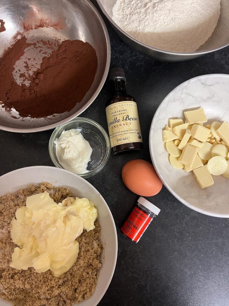 bowls of flour, cocoa powder, brown sugar, butter, white chocolate chunks, a bottle of vanilla extract, an egg and a red tub of food colouring.