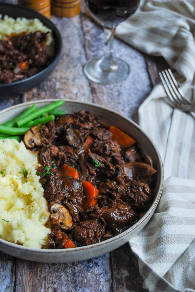 a bowl of beef Bourguignon with carrots, mushrooms and shallots with mashed potatoes and green beans, a glass of red wine and a striped tea towel.