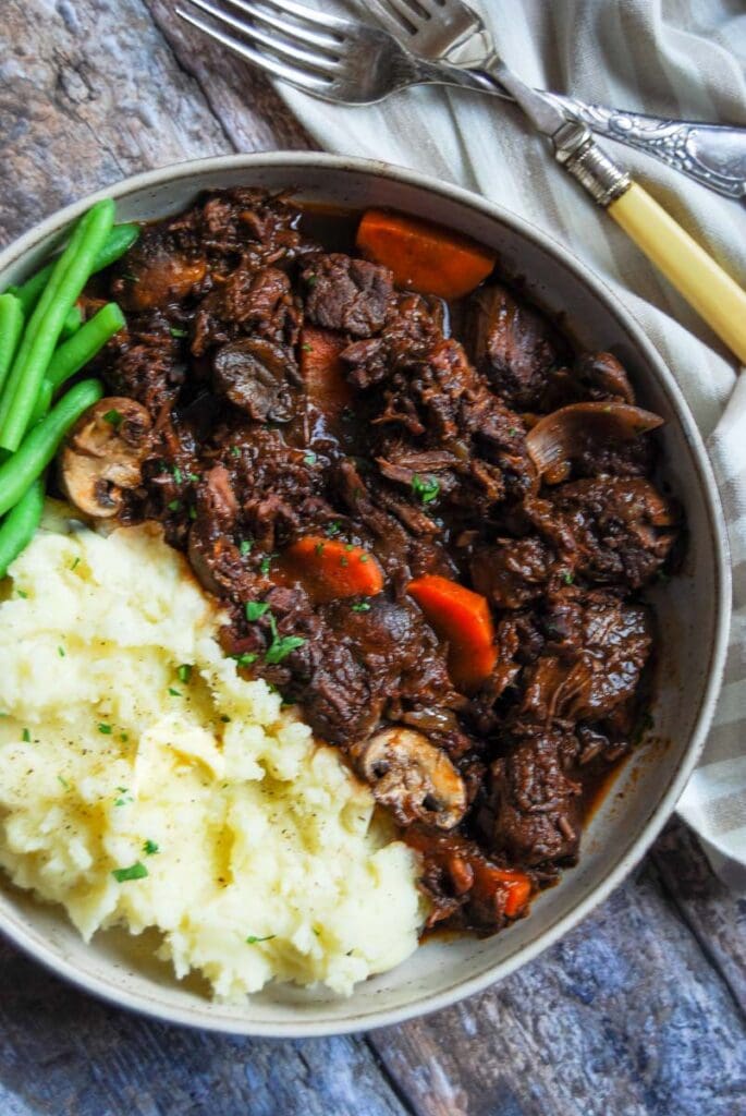 a bowl of beef stew in red wine gravy with carrots, mushrooms and shallots with mashed potatoes and green beans beside a beige and white tea towel and serving fork. 