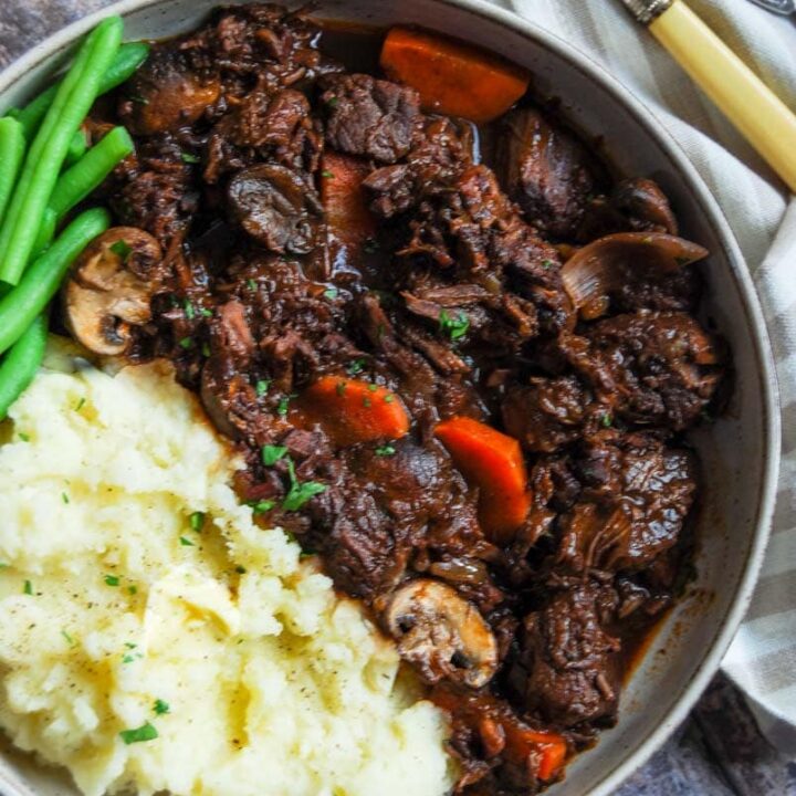 a bowl of beef stew in red wine gravy with carrots, mushrooms and shallots with mashed potatoes and green beans beside a beige and white tea towel and serving fork.