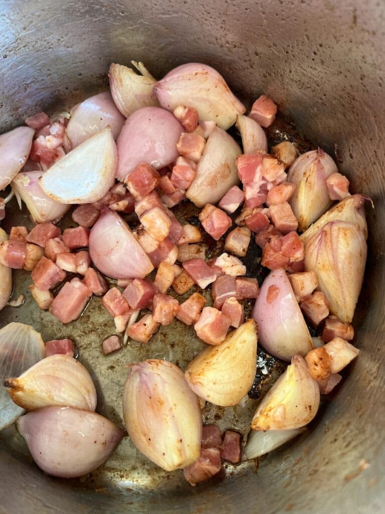 bacon lardons and peeled halved shallots cooking in a silver pan.