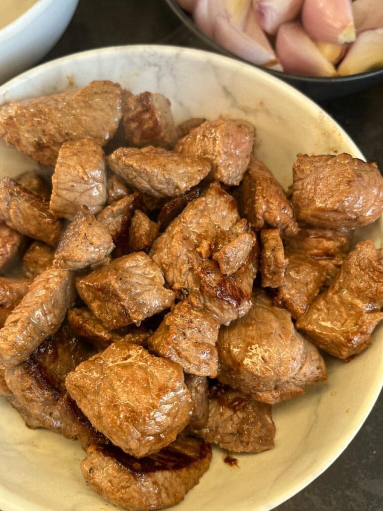 a white bowl of browned beef chunks and a bowl of peeled shallots.