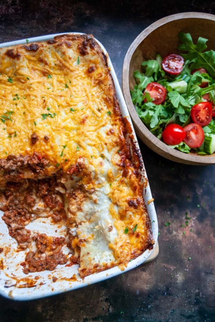 a homemade lasagne topped with cheese and a wooden bowl of green salad and tomatoes.