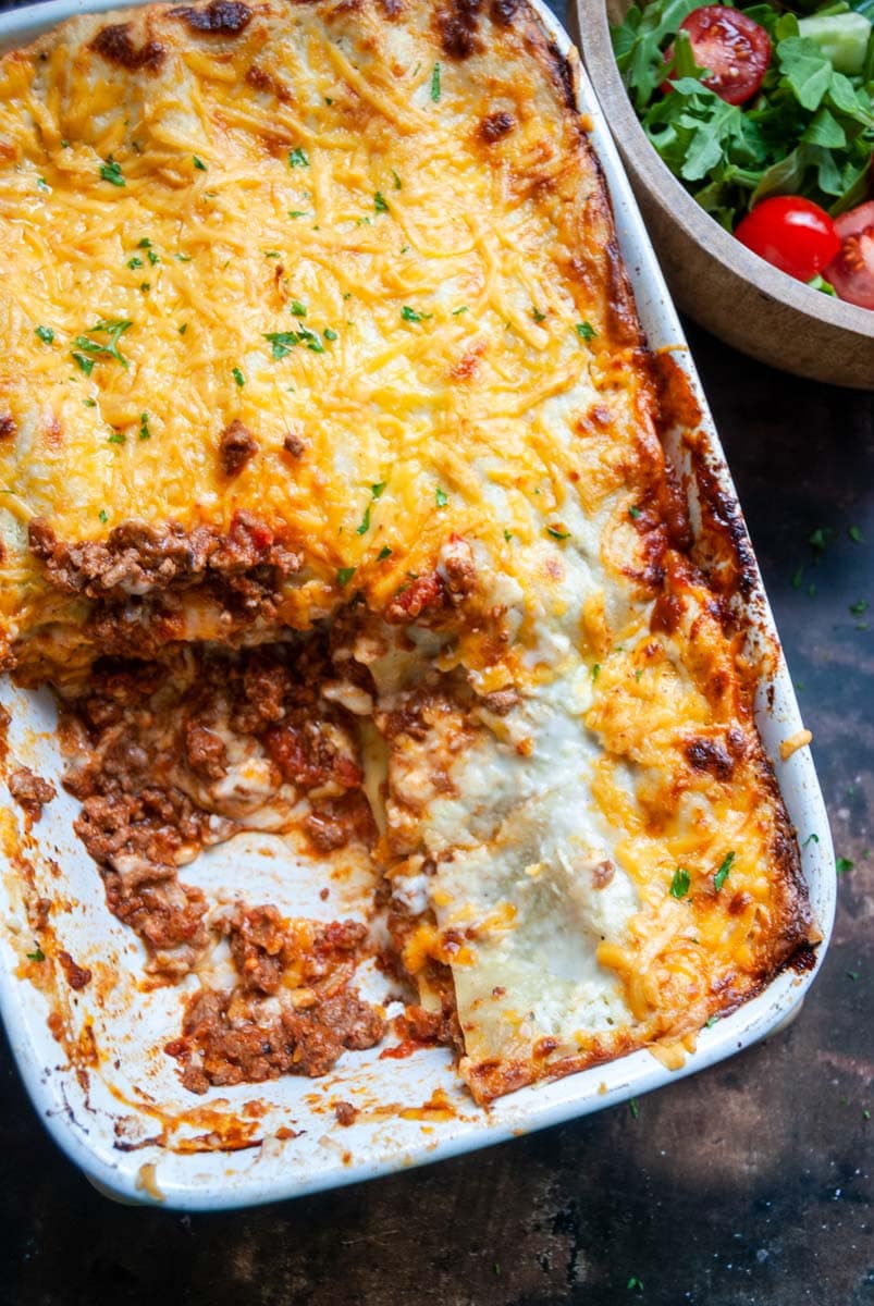 A large lasagne topped with cheese in a white dish and a wooden bowl of green salad and tomatoes.