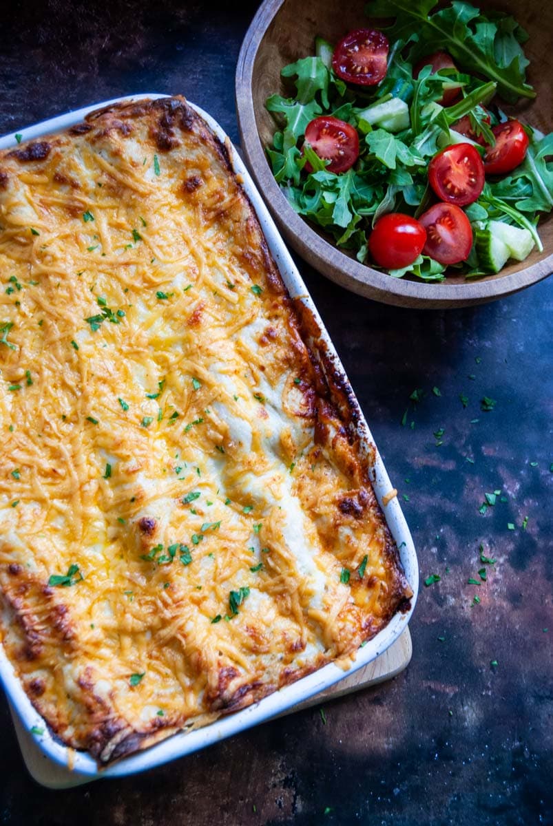 a homemade lasagne in a white dish with a wooden bowl of lettuce and tomatoes.