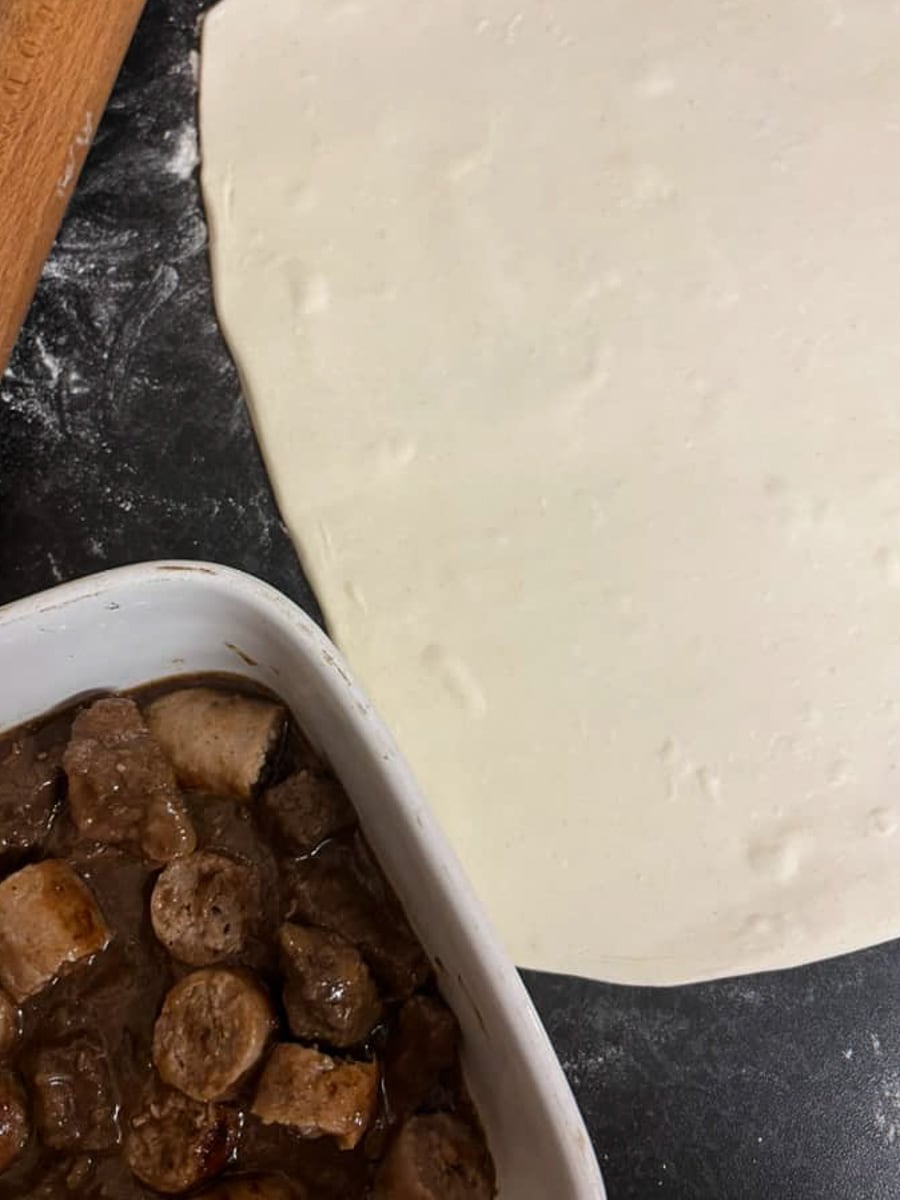 a rolled out block of puff pastry, a rolling pin and a white dish of beef and sausage stew.