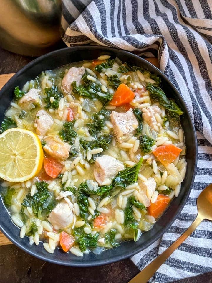 a bowl of chicken vegetable soup with orzo pasta and a lemon slice, a striped tea towel and a gold spoon.