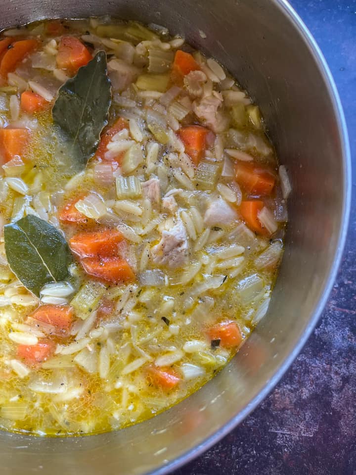 a silver pan of chicken soup with orzo pasta and bay leaves.