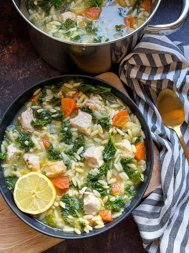 a black bowl of chicken soup with carrots, kale, leeks and orzo pasta with a slice of lemon, a striped tea towel, a gold spoon and a large pan of soup.