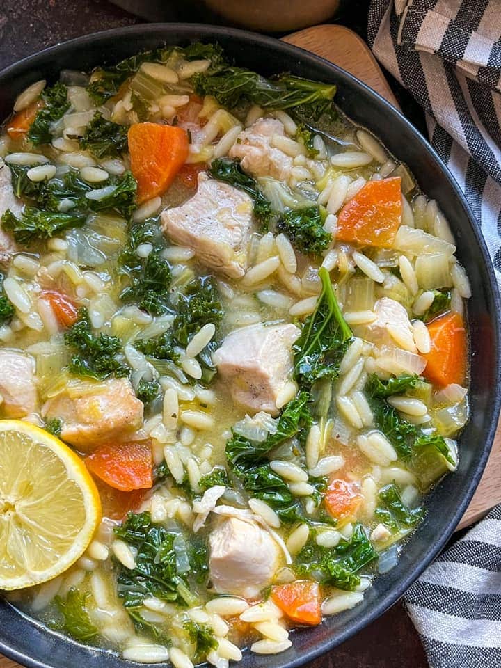 a bowl of chicken soup with orzo pasta, carrots, kale and leeks with a lemon slice.