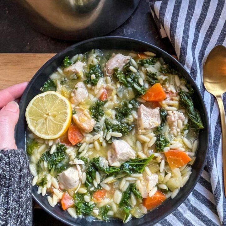 a black stoneware bowl of chicken soup with orzo pasta, kale, carrots and a slice of lemon, a striped tea towel and a gold spoon.