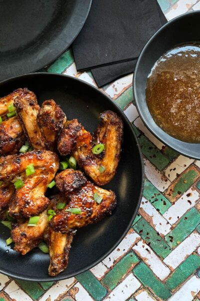 a black stoneware bowl of chicken wings sprinkled with chopped green onins, a bowl of hot honey sauce and black paper napkins.