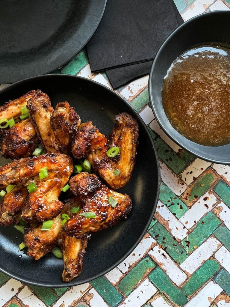 a black stoneware bowl of chicken wings sprinkled with chopped green onins, a bowl of hot honey sauce and black paper napkins.