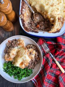 A plate of haggis, neeps and tatties with steamed kale, wooden salt and pepper mills, a tartan tea towel, a silver fork with a yellow handle and a pie dish of the haggis, neeps and tatties.