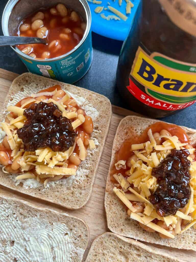 two slices of buttered brown bread topped with baked beans, grated cheese and pickle, a small can of baked beans and a jar of pickle.