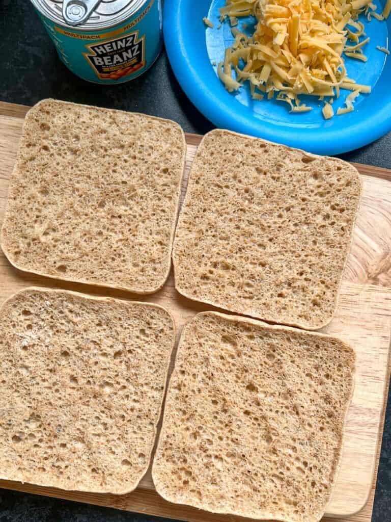 a wooden chopping board with 4 slices of brown bread, a blue plate of grated cheese and a small tin of baked beans.