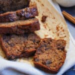 a partially sliced banana bread on a wire cooling rack covered with a beige tea towel, cinnamon sticks and a speckled beige and white coffee mug.