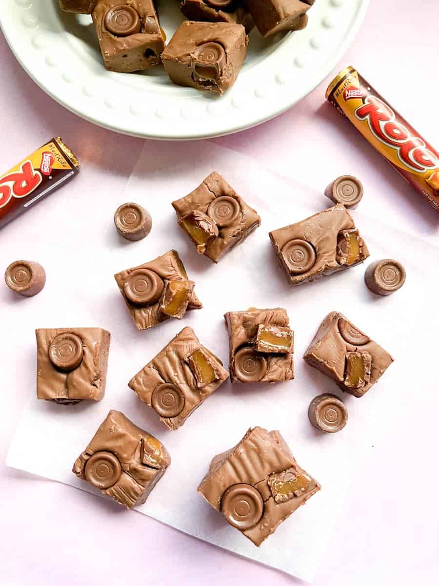 nine pieces of Rolo fudge on a piece of baking paper, two tubes of Rolo sweets and a white plate of fudge.