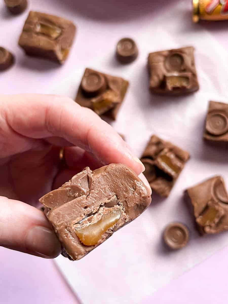 a hand holding a piece of chocolate Rolo fudge with more fudge and Rolo sweets in the background.