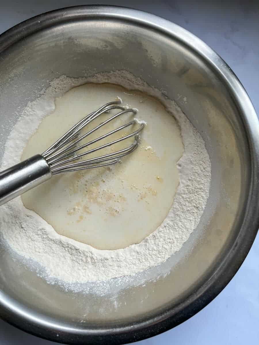 a silver mixing bowl of flour, eggs and milk and a silver whisk.