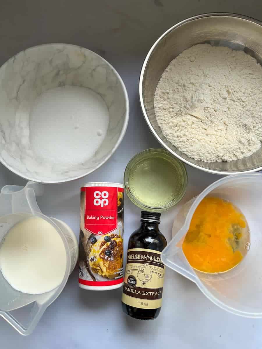 silver bowls of flour, sugar, a jug of milk, a jug of beaten eggs, a small jar of oil, a tub of baking powder and a bottle of vanilla extract.