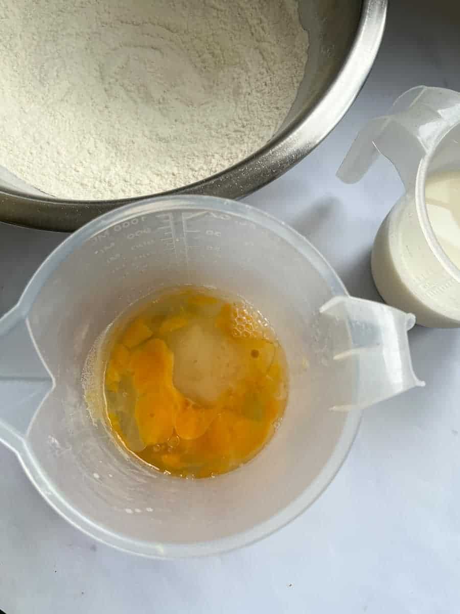 a small jug of beaten eggs and oil, a large silver mixing bowl of flour and a small jug of milk.
