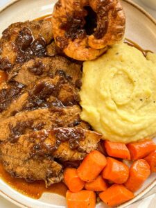 a plate of sliced cooked beef in onion gravy with mashed potatoes and carrots.